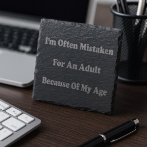 Laser-engraved slate plaque with the quote ‘I’m Often Mistaken For An Adult Because Of My Age’ displayed on an office desk beside a laptop, keyboard, pen and stationery holder