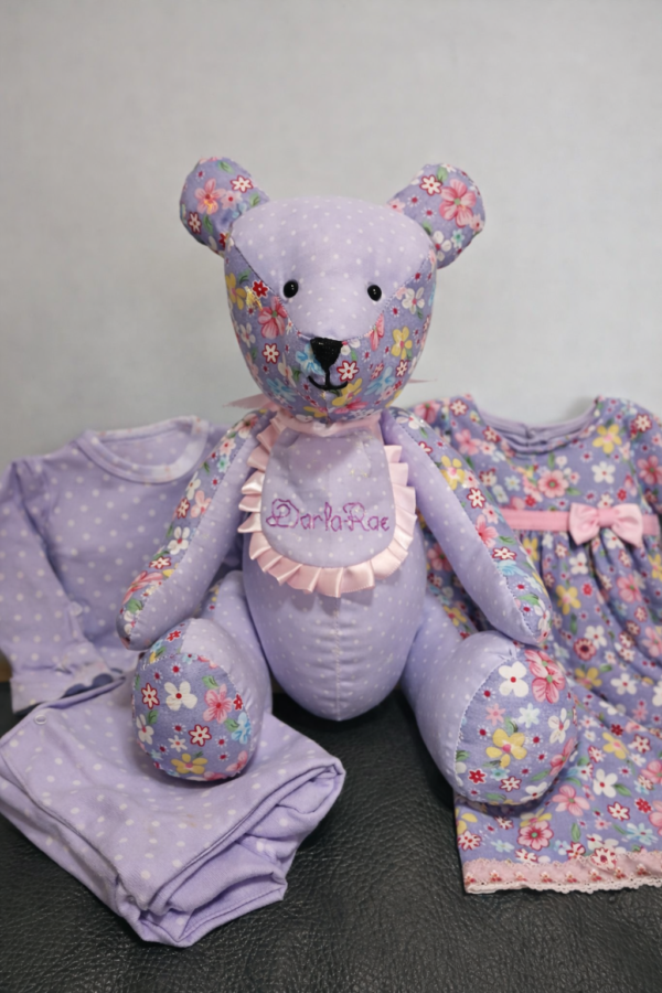 Handmade memory bear created from baby clothing, displayed with the original garments used in its creation, featuring soft lilac floral fabrics.