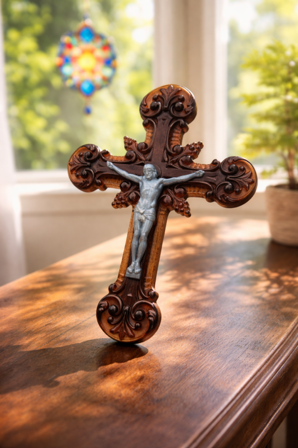 Ornate acrylic crucifix with silver Christ figure displayed on a polished table in soft window sunlight with stained glass suncatcher in background