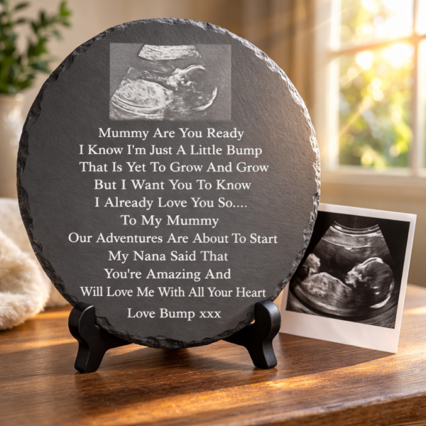 Round slate plaque engraved with an ultrasound baby scan and pregnancy poem displayed on a stand beside a baby scan photo in warm sunlight.