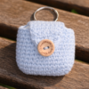 Light blue handmade crocheted AirPods case with keyring and wooden button displayed on a sunny wooden park bench